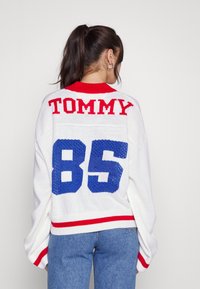 Bílý pletený svetr s červenými akcenty, na zadní straně s nápisem "TOMMY" červenými písmeny a "85" modře. Volný, pohodlný střih s žebrovaným lemem.