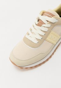 Beige treningssko med mesh- og semsket skinnmaterialer, kremfargede lisser, brune detaljer, teksturert såle og en "BORG"-logo på siden.