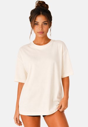 Junge Frau mit zusammengebundenen Haaren, die ein lockeres, schlichtes weißes T-Shirt und schwarze Shorts trägt, steht vor einem einfarbigen hellen Hintergrund.