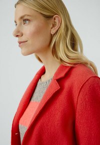 Manteau en mélange de laine rouge avec une finition texturée, revers crantés et coupe ajustée. Superposé sur un pull gris avec une rayure rose.