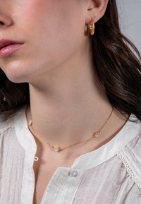 Collana d'oro con accenti floreali e piccoli cristalli, abbinata a orecchini a cerchio d'oro, indossata su un top in tessuto leggero e testurizzato.