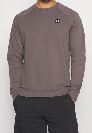 Man wearing a mauve long-sleeve crewneck sweatshirt with a small black and white logo on the chest and black pants, hands in pockets.