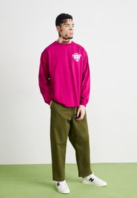 Långärmad rosa T-shirt med en vit grafisk logotyp, kombinerad med lösa olivgröna byxor och vita sneakers med svarta detaljer.