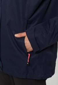 Main à peau claire dans la poche d'une veste bleu foncé avec un curseur de fermeture éclair rose et un poignet en velcro.
