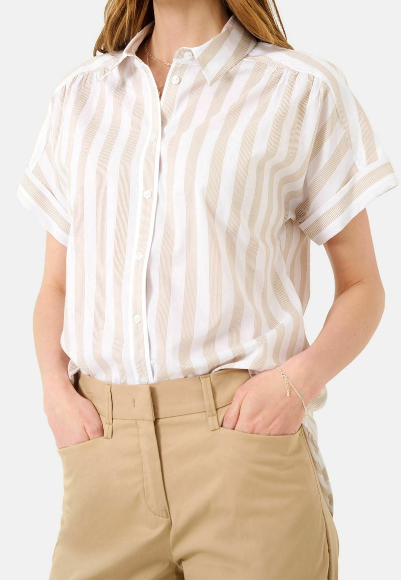 Femme portant une chemise à boutons à manches courtes rayée verticalement beige et blanche, rentrée dans un pantalon beige, mains dans les poches.