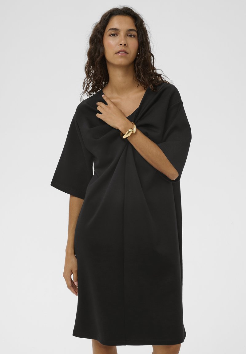 Vestido negro de gran tamaño con mangas cortas, que presenta un escote en V profundo y detalles drapeados en la parte delantera. Textura de tela suave, largo hasta la rodilla.