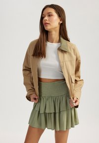 Veste tan avec col vert, associée à une jupe verte à plusieurs couches avec une taille smockée. Crop top blanc en dessous.