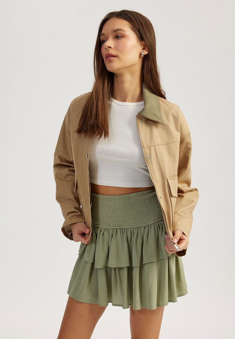 Veste tan avec col vert, associée à une jupe verte à plusieurs couches avec une taille smockée. Crop top blanc en dessous.