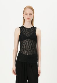 Svart blonde tanktop med en gjennomsiktig bodice, med intrikate diamantmønstre og solid svart kant. Modellen bruker den med løse svarte bukser.