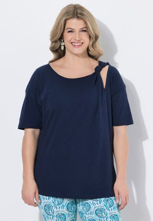 Mujer sonriente con cabello rubio ondulado lleva un top asimétrico azul marino con detalle de nudo y pantalones azules con estampado, sobre fondo blanco.