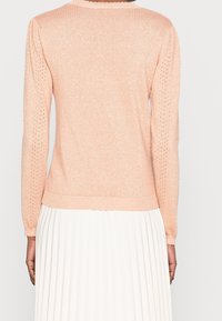 Pull léger de couleur pêche avec des manches longues, col côtelé et détails en tricot ouvert texturé le long des manches. Tissu doux et scintillant.