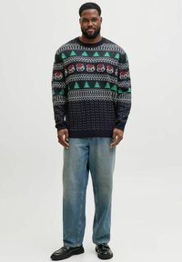 Brede trui met een marineblauwe basis, groene bomen, rode cadeaus en witte patronen. Gepaard met lichtblauwe denim en zwarte schoenen.