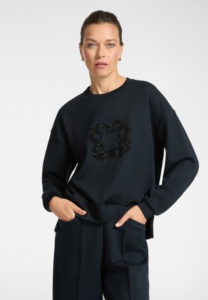Donna con i capelli raccolti, che indossa una felpa navy scuro con un motivo floreale di paillettes nere e pantaloni abbinati, mani in tasca, sfondo neutro.