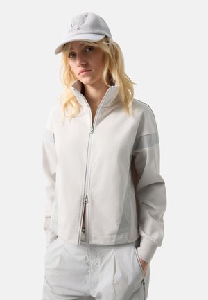 Młoda kobieta z blond włosami, ubrana w jasną kurtkę zapinaną na zamek, pasujące spodnie i białą czapkę z daszkiem, stoi z rękami w kieszeniach.