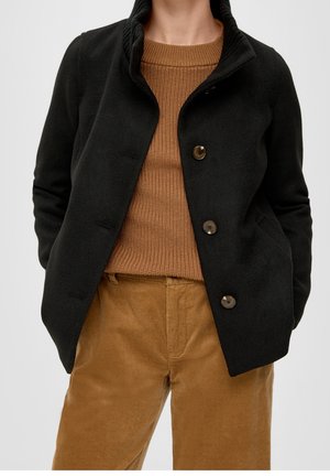 Osoba s černou zapínací bundou přes hnědý pletený svetr a hnědé manšestrové kalhoty, stojící s rukama v kapsách.