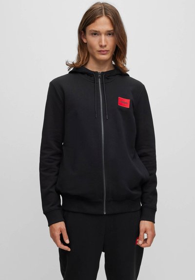 Jeune personne aux cheveux mi-longs porte un sweat à capuche noir zippé avec un patch logo rouge et un pantalon noir assorti sur fond uni.