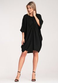 Czarna, oversize sukienka wykonana z miękkiego materiału, z detalem na środku w postaci węzła, krótkimi rękawami oraz luźnym, swobodnym krojem.