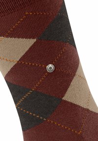 Calcetín con patrón argyle en marrón, beige y gris oscuro, y pequeño logotipo metálico redondo en la tela.