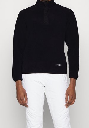Personne portant un pull noir à manches longues avec un col haut et un pantalon blanc, debout devant un fond uni.