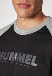 Svart T-skjorte med grå raglan-ermer, med en dristig trykket logo "HUMMEL" og teksten "sportswear since 1923" i kontrastfarger.