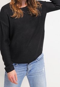 Černý pletený svetr s volným střihem, žebrovanými manžetami a lemem. Nošený s světle modrými roztrhanými džínami, představujícími ležérní design.