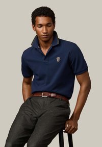 Granatowa koszulka polo z kontrastującym zielonym kołnierzykiem w paski i haftowanym logo. Noszona z szarymi spodniami o teksturowanej strukturze oraz plecionym brązowym paskiem.