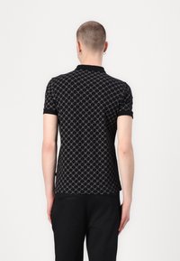 Personne aux cheveux courts portant un polo noir à manches courtes à motif et un pantalon noir, debout de dos sur un fond uni.