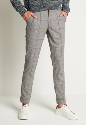 Personne portant un pantalon slim à carreaux gris, les mains dans les poches, avec des baskets blanches, debout devant un fond neutre.