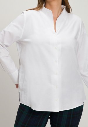 Personne portant une chemise blanche à boutons avec un col montant et un pantalon à carreaux vert foncé, debout avec une main sur la hanche.