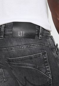 Parche de cuero negro con el logo "LTB" en la cinta de la cintura de unos vaqueros de denim gris, con un leve desgaste y detalles de costura en contraste.
