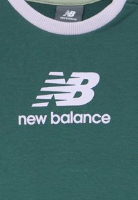 Πράσινο μπλουζάκι με στρογγυλή λαιμόκοψη και λευκή ribbed περιφέρεια, που εμφανίζει μεγάλο λευκό λογότυπο New Balance και κείμενο κεντραρισμένο στο μπροστινό μέρος.