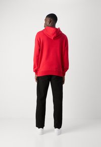 Rød hættetrøje med afslappet pasform, ribbede manchetter og kant, parret med sorte lige ben bukser og hvide sneakers på en neutral baggrund.