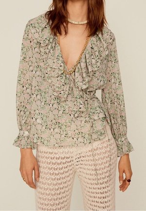 Kvinde iført en blomsterprintet bluse med flæser og dyb V-hals samt hørbukser med hæklet mønster, accessoriseret med halskæder med træperler.