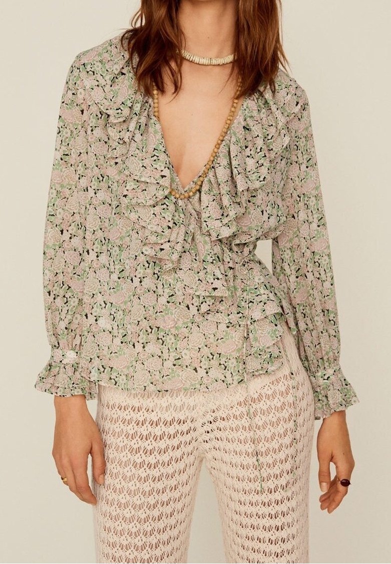 Femme portant un chemisier à volants fleuri avec un décolleté en V profond et un pantalon beige à motif crocheté, accessoirisée de colliers en perles de bois.