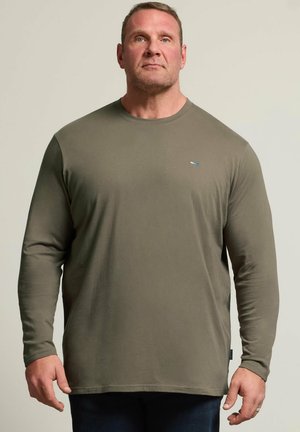 Mand med kort hår iført en olivengrøn langærmet skjorte, stående foran en ensfarvet lys baggrund, vendt mod fronten.