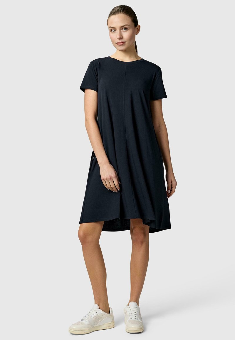 Femme debout portant une robe noire ample à manches courtes et des baskets blanches, sur un fond gris clair uni.