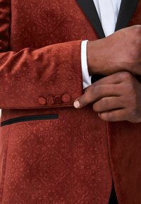 Strukturierter rostfarbener Blazer mit subtilen Mustern, schwarzer Revers und vier mit Stoff bezogenen Knöpfen am Ärmel. Schwarzes Taschendetail sichtbar.