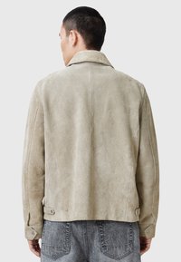 Beige mockasinsjacka med bred krage, synlig sömnad och sidoknappar. Texturerad yta, strukturerad passform och medellång design.