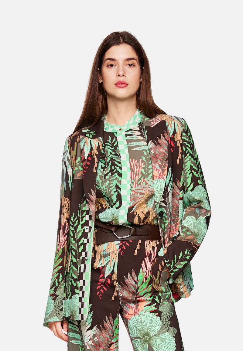 Multicolorierter Blazer und Oberteil mit Blumenmuster und grünen Karos auf braunem Hintergrund, kombiniert mit passender Hose und einem braunen Gürtel.