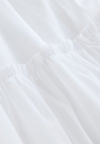 Tela blanca con cintura elástica fruncida. Textura translúcida con sutiles detalles de patrón, creando suaves pliegues y caídas.