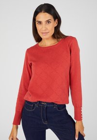 Rode gebreide trui met een ruitpatroon, ronde halslijn en knoopaccenten op de mouwen, gecombineerd met donkerblauwe jeans.