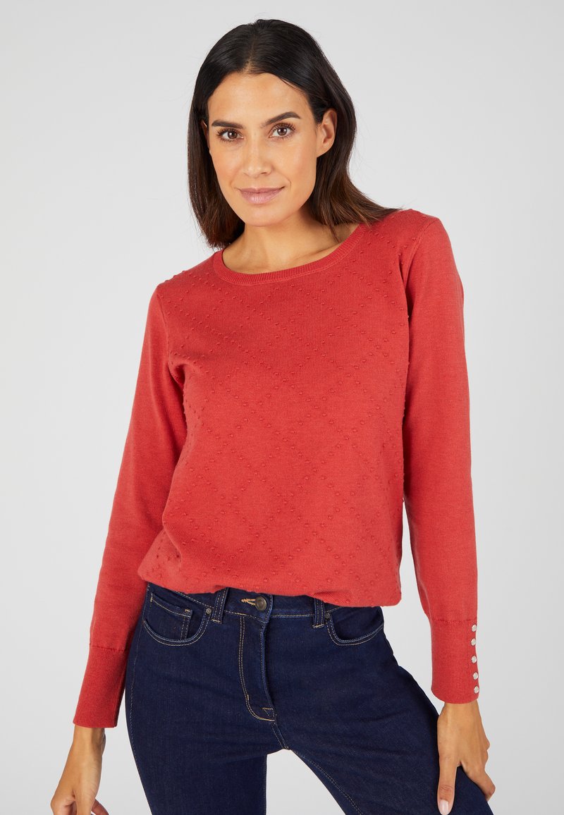 Rode gebreide trui met een ruitpatroon, ronde halslijn en knoopaccenten op de mouwen, gecombineerd met donkerblauwe jeans.