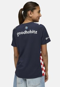 Mornarica sportska majica s bočnim panelima u crveno-bijelim kockicama. Sadrži "goodhabitz" logo i grb na leđima, kratke rukave i prozračnu tkaninu.