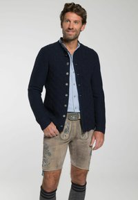Marine gebreide cardigan met een textuurpatroon, zilveren knopen, gedragen over een lichtblauw shirt en beige shorts met blauwe borduursels en donkergrijze sokken.