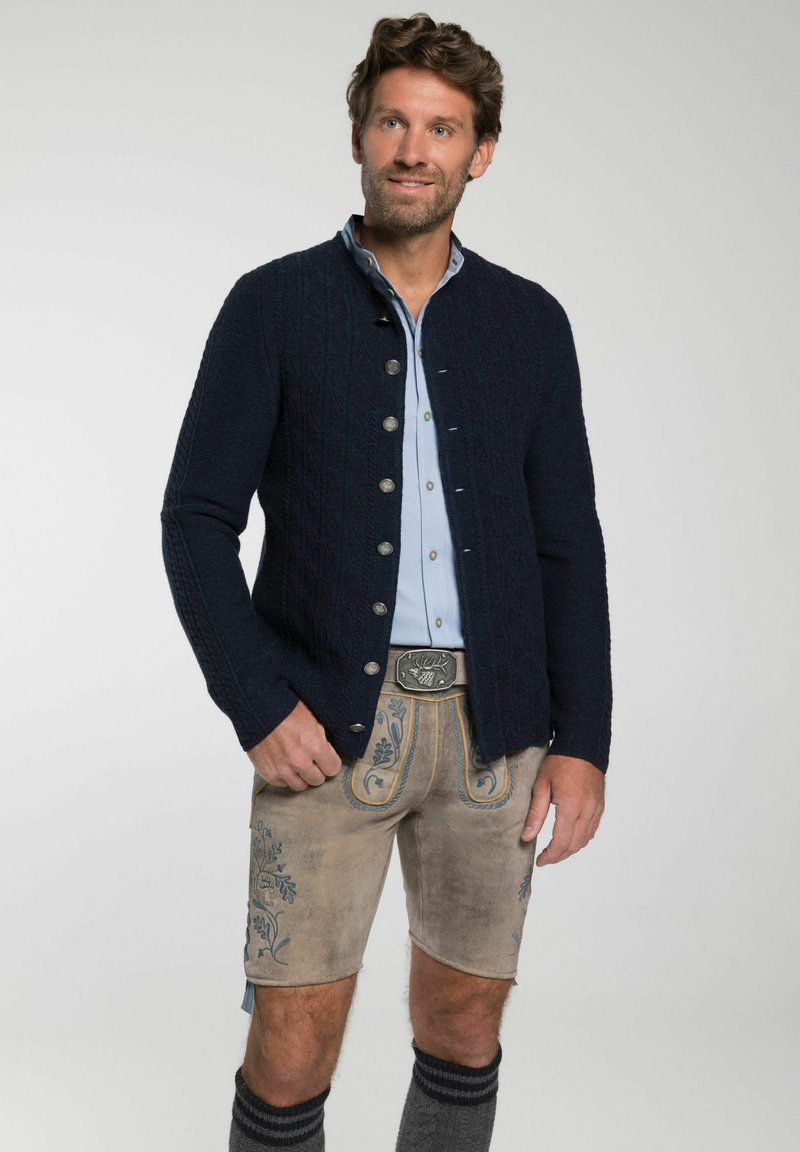 Marine gebreide cardigan met een textuurpatroon, zilveren knopen, gedragen over een lichtblauw shirt en beige shorts met blauwe borduursels en donkergrijze sokken.