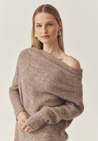 Eine Frau mit langen blonden Haaren trägt einen schulterfreien, weichen, beigen Pullover und große goldene Ohrringe, während sie vor einem schlichten, hellen Hintergrund steht.