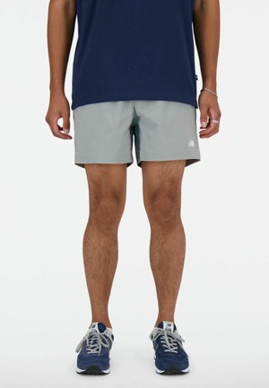 Homme portant un short gris, un t-shirt bleu marine et des baskets bleues et blanches, debout les bras détendus le long du corps.