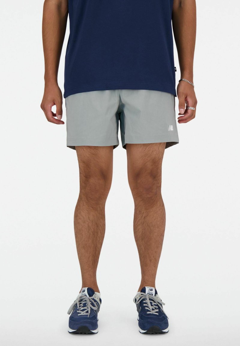Homme portant un short gris, un t-shirt bleu marine et des baskets bleues et blanches, debout les bras détendus le long du corps.