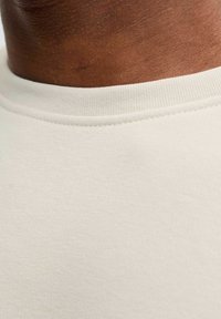 T-shirt beige clair avec une texture douce et lisse, et un col rond simple. Le tissu semble extensible et léger.