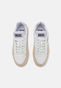 Des baskets en cuir blanc présentent un design perforé sur l'extrémité, des lacets plats blancs et une semelle en caoutchouc beige. Marquage sur la languette.